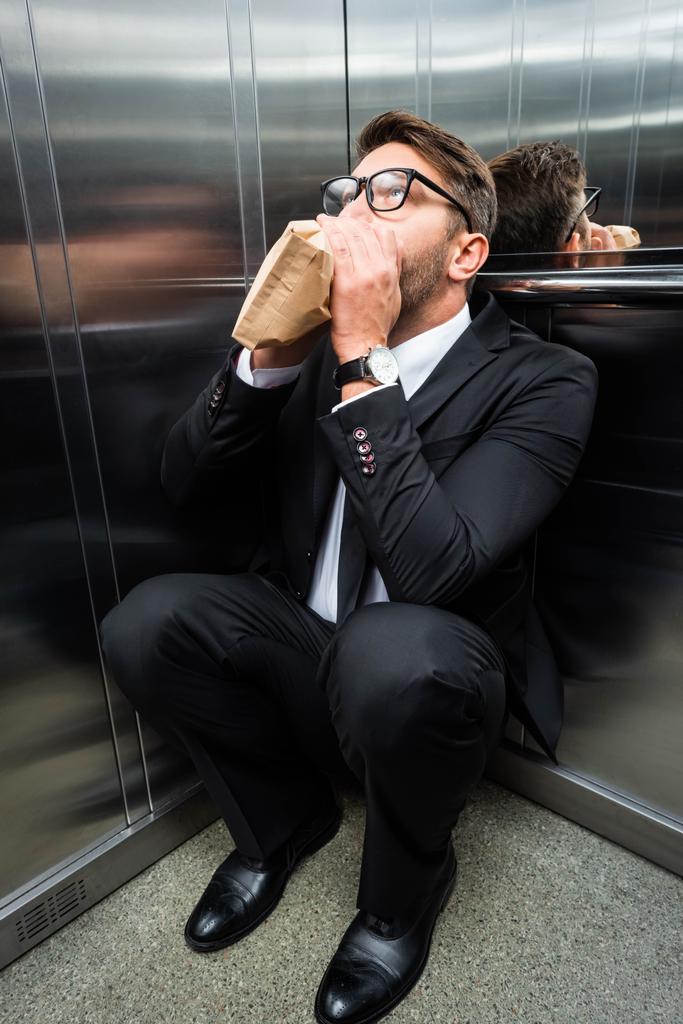 испуганный бизнесмен в костюме с клаустрофобией дышит бумажным пакетом в лифте
 - Фото, изображение
