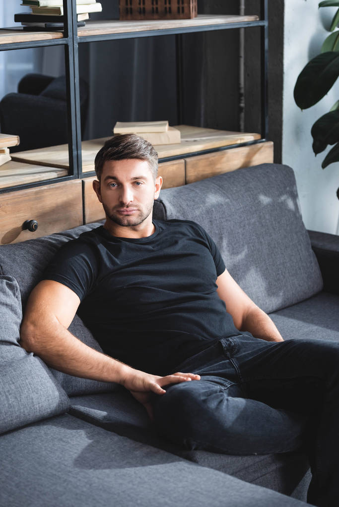 przystojny mężczyzna w czarny t-shirt siedzi na kanapie w mieszkaniu  - Zdjęcie, obraz
