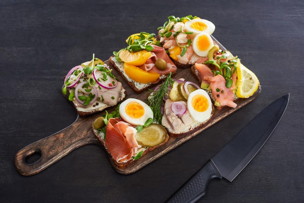 Holzschneidebrett mit dänischen Smorrebrod-Sandwiches in Messernähe auf grauer Oberfläche  - Foto, Bild