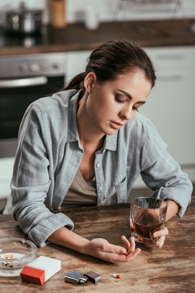 Femme inquiète tenant verre de whisky et pilules à la table de la cuisine
 - Photo, image