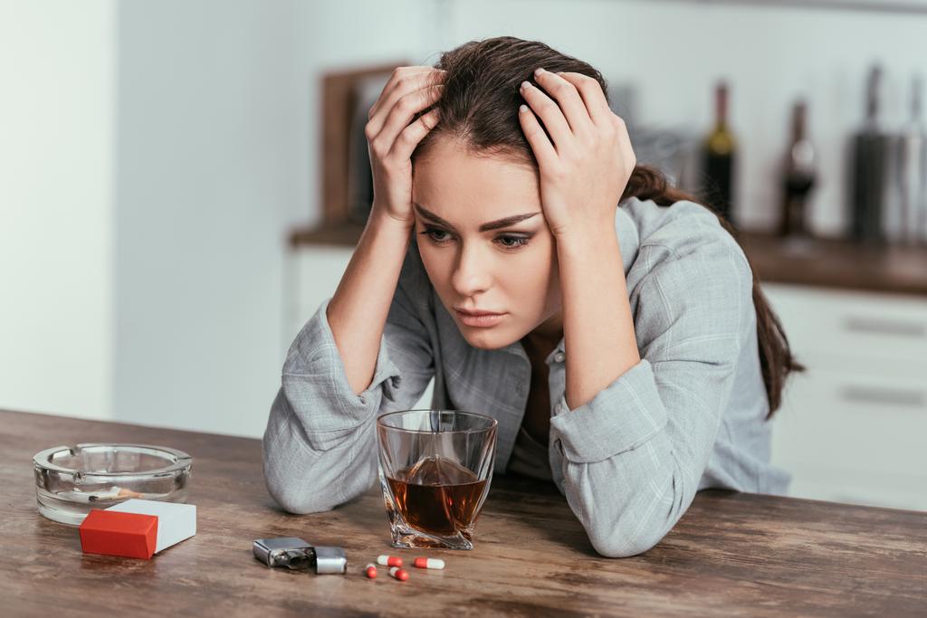 Rozczarowana kobieta siedząca przy stole z whisky, tabletkami i papierosami - Zdjęcie, obraz