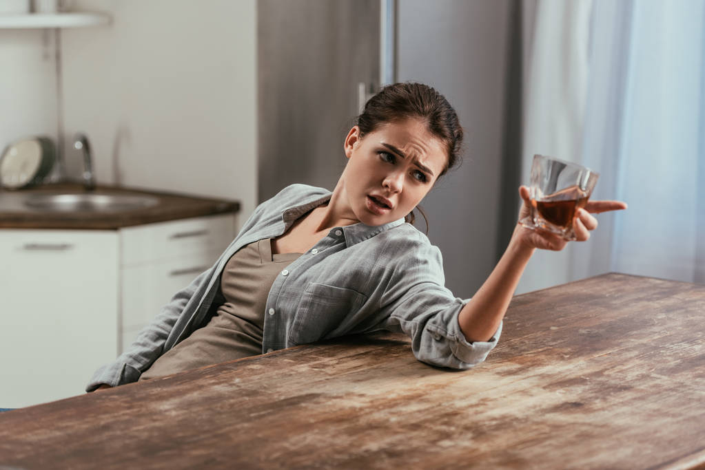 Angry woman with whiskey glass talking to somewhere at kitchen table - 写真・画像