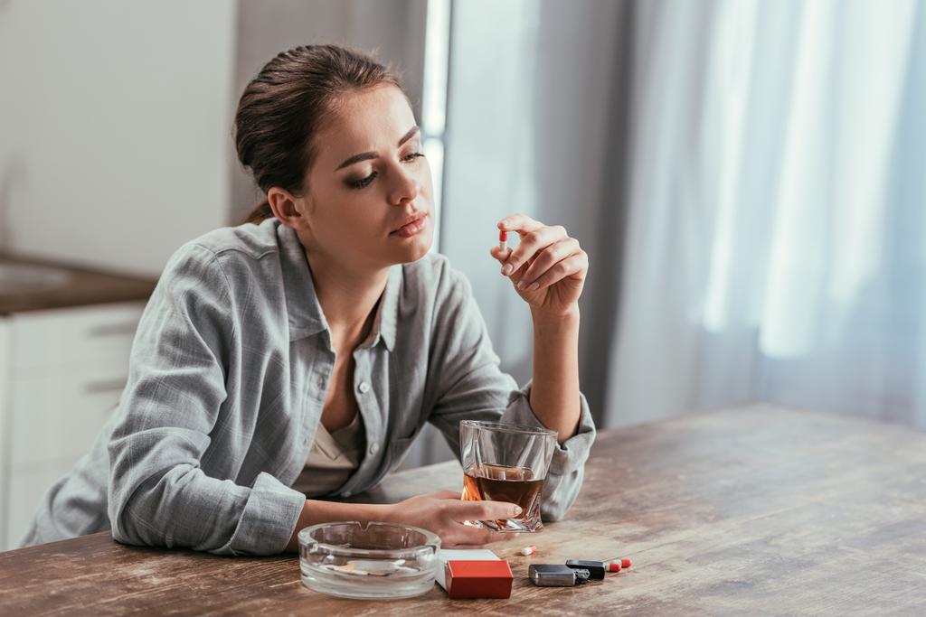 Mulher viciada em álcool segurando pílula e copo de uísque ao lado de cigarros na mesa
 - Foto, Imagem