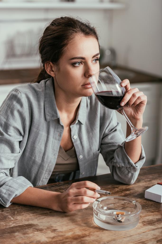 Töprengő nő bort iszik és dohányzik a konyhaasztalnál - Fotó, kép