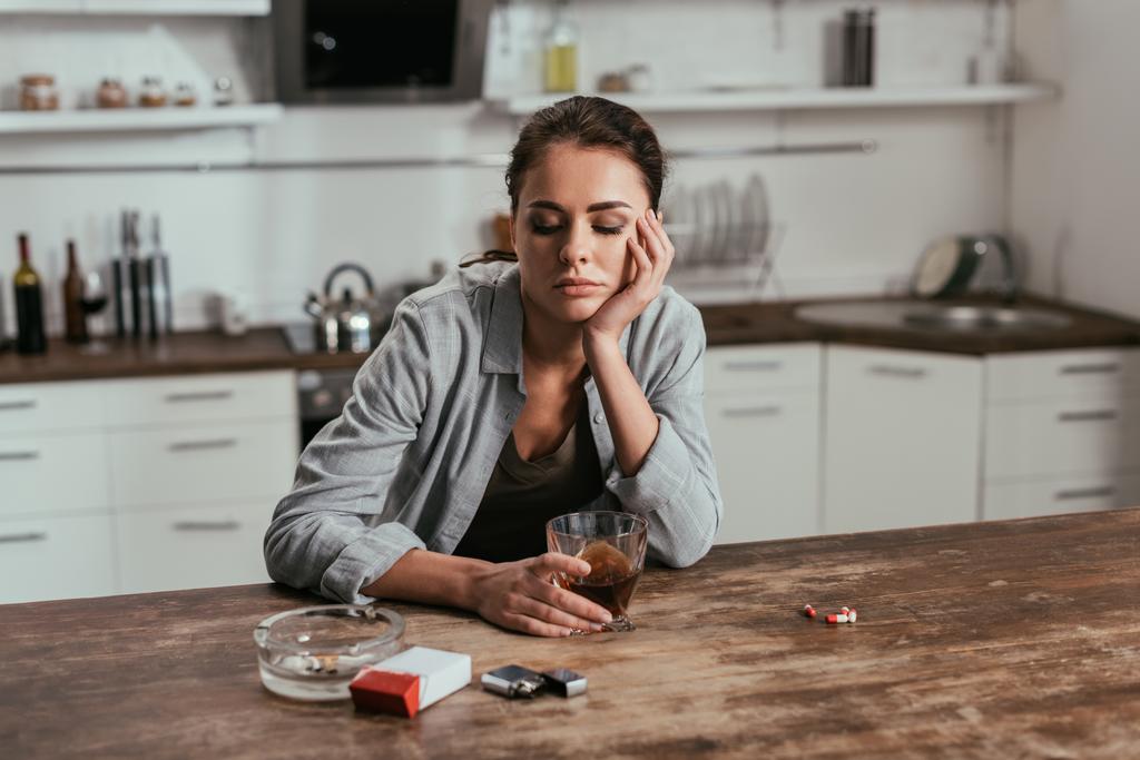 Huolestunut nainen pitelee viskilasia savukkeiden ja pillereiden vieressä pöydällä
  - Valokuva, kuva