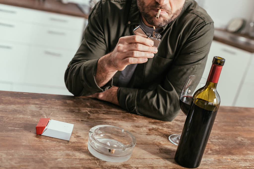 Gewassen beeld van een man die naast wijnfles en glas op tafel een sigaret aansteekt - Foto, afbeelding