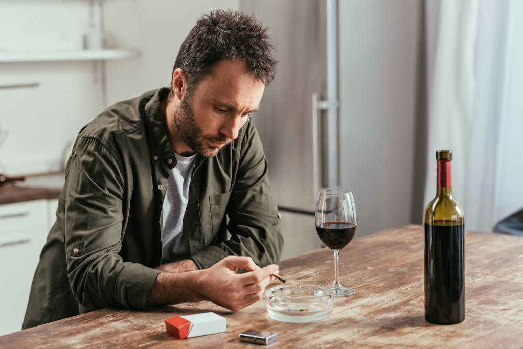 Розчарований чоловік курить сигарету поруч з пляшкою вина та склом на кухонному столі
 - Фото, зображення