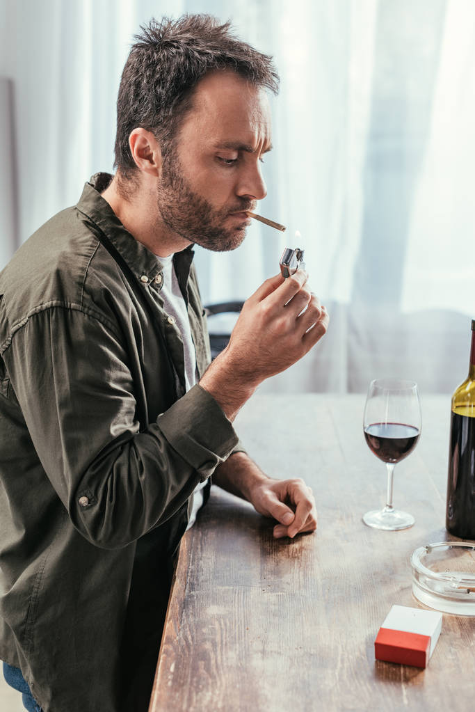 Человек, зажигающий сигарету рядом с бутылкой вина и бокалом на столе
 - Фото, изображение