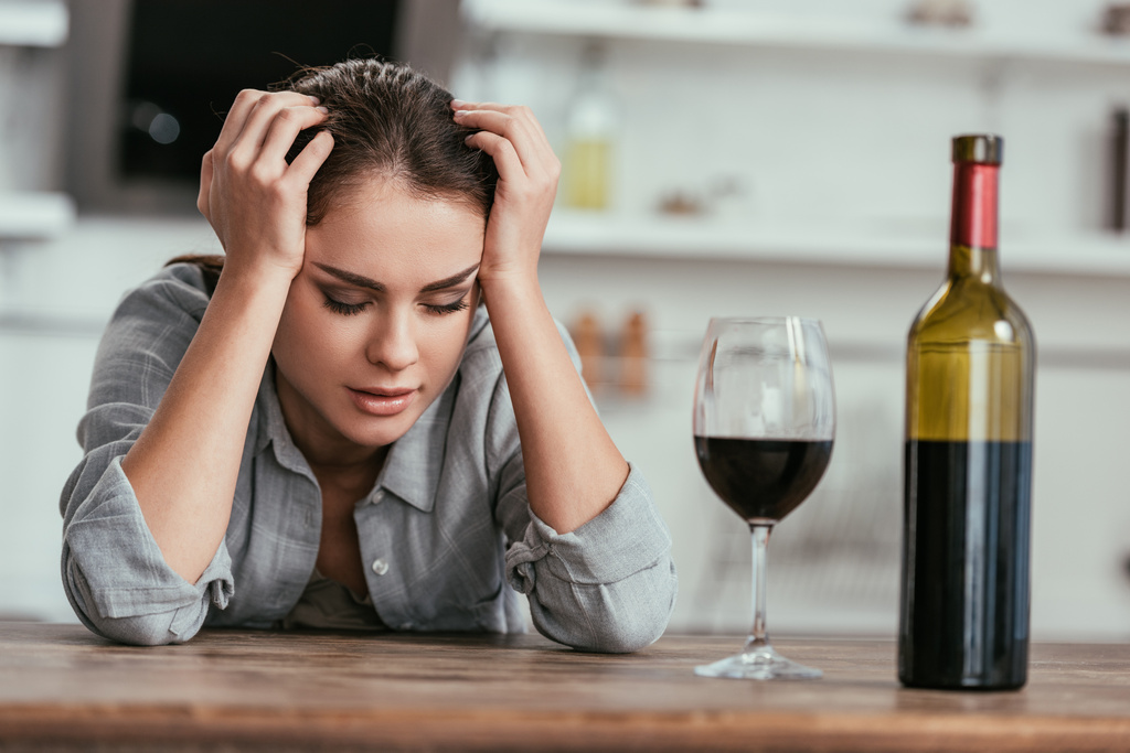 Selektiver Fokus einer alkoholabhängigen Frau, die neben Wein am Küchentisch sitzt - Foto, Bild