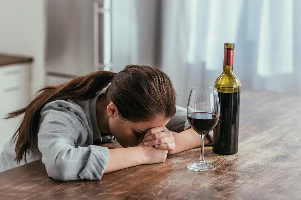 Απογοητευμένη γυναίκα δίπλα σε ποτήρι κρασιού και μπουκάλι στο τραπέζι - Φωτογραφία, εικόνα