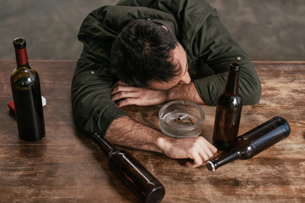 Высокий угол обзора пьяного мужчины лежащего на столе с бутылками и пепельницей
 - Фото, изображение