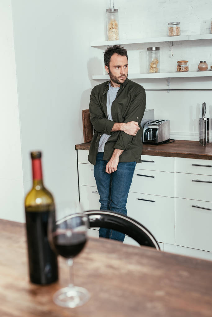 Selective focus of sad man standing on kitchen and wine glass with bottle on table - 写真・画像