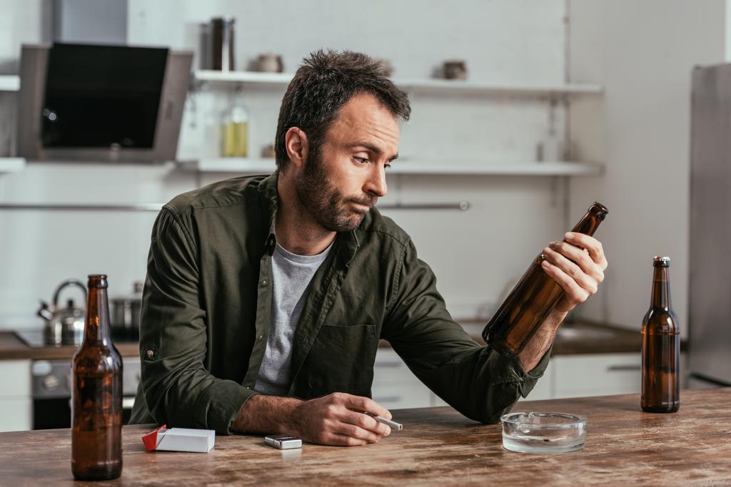 Λυπημένος άνθρωπος με αλκοόλ εξαρτάται κρατώντας τσιγάρο και μπύρα μπουκάλι στο τραπέζι - Φωτογραφία, εικόνα