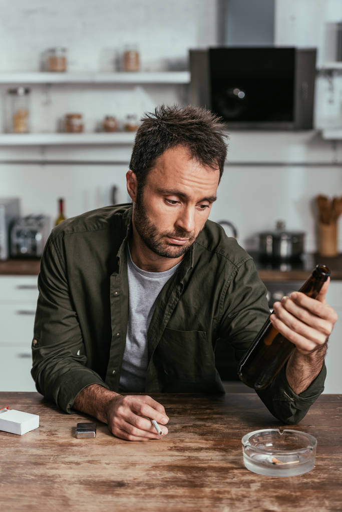 Rozrušený muž s alkoholem spoléhal na to, že drží u stolu cigaretu a láhev od piva - Fotografie, Obrázek