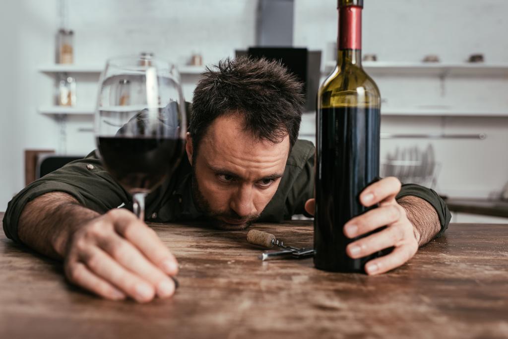 Mutfak masasında şarap kadehi ve şişe tutan üzgün adamın seçici odak noktası. - Fotoğraf, Görsel