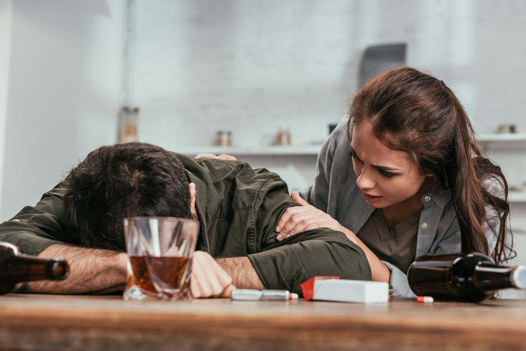 Επιλεκτική εστίαση της γυναίκας που μιλάει με μεθυσμένο σύζυγο δίπλα σε μπουκάλια και τσιγάρα στο τραπέζι - Φωτογραφία, εικόνα