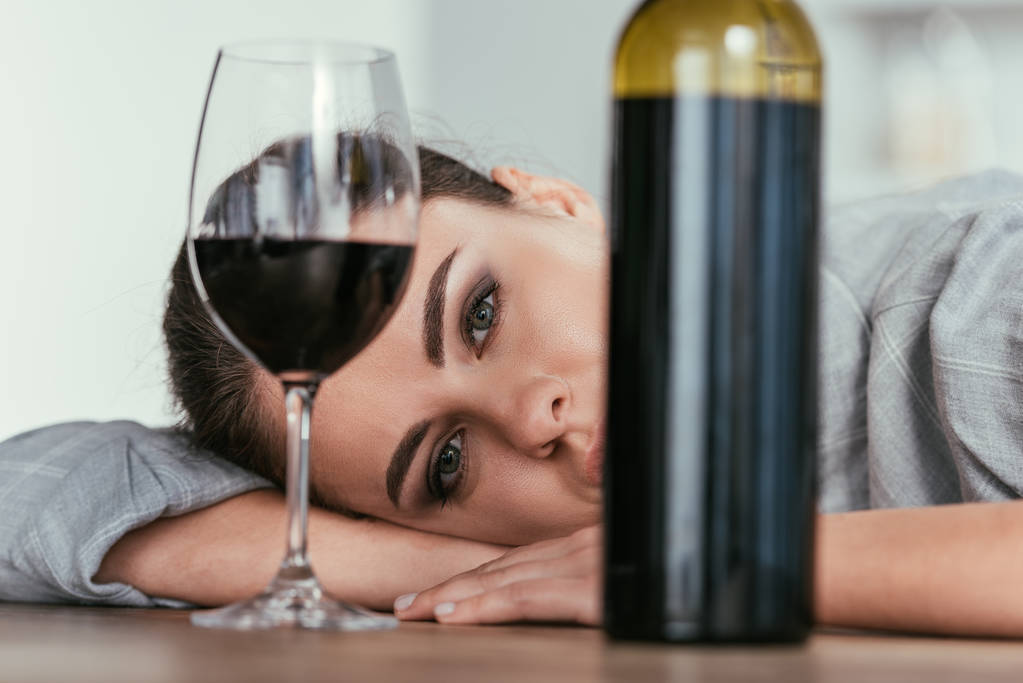 Селективный фокус женщины, смотрящей в камеру с бокалом вина и бутылкой на столе
 - Фото, изображение