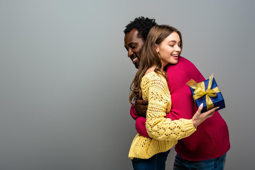 Ευτυχισμένο διαφυλετικό ζευγάρι με πουλόβερ που κρατάει το παρόν και αγκαλιάζει σε γκρι φόντο - Φωτογραφία, εικόνα