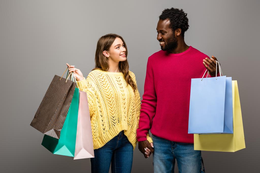 szczęśliwa międzyrasowa para w swetrach trzymająca się za ręce i torby na zakupy na szarym tle - Zdjęcie, obraz
