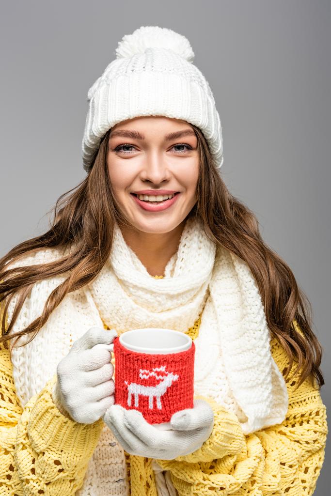 happy girl in yellow sweater, scarf, hat and gloves holding mug of cocoa isolated on grey - Photo, Image