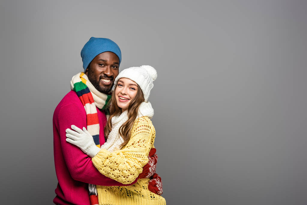 χαμογελαστό διαφυλετικό ζευγάρι με χειμωνιάτικη στολή αγκαλιασμένο σε γκρι φόντο - Φωτογραφία, εικόνα