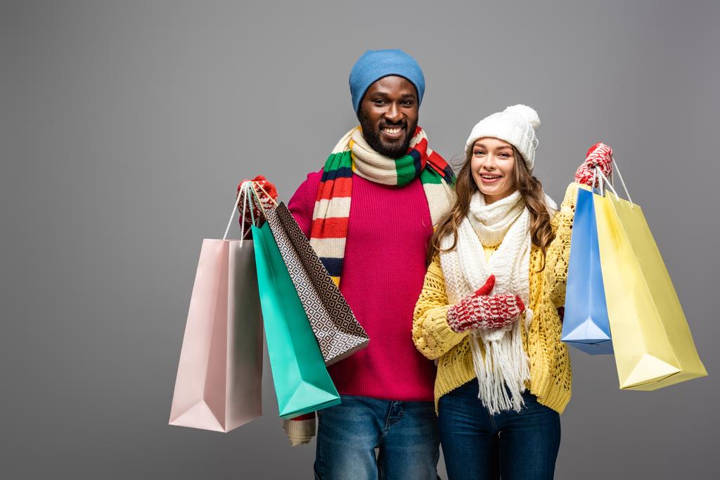 feliz pareja interracial en invierno traje celebración bolsas de compras sobre fondo gris
 - Foto, Imagen