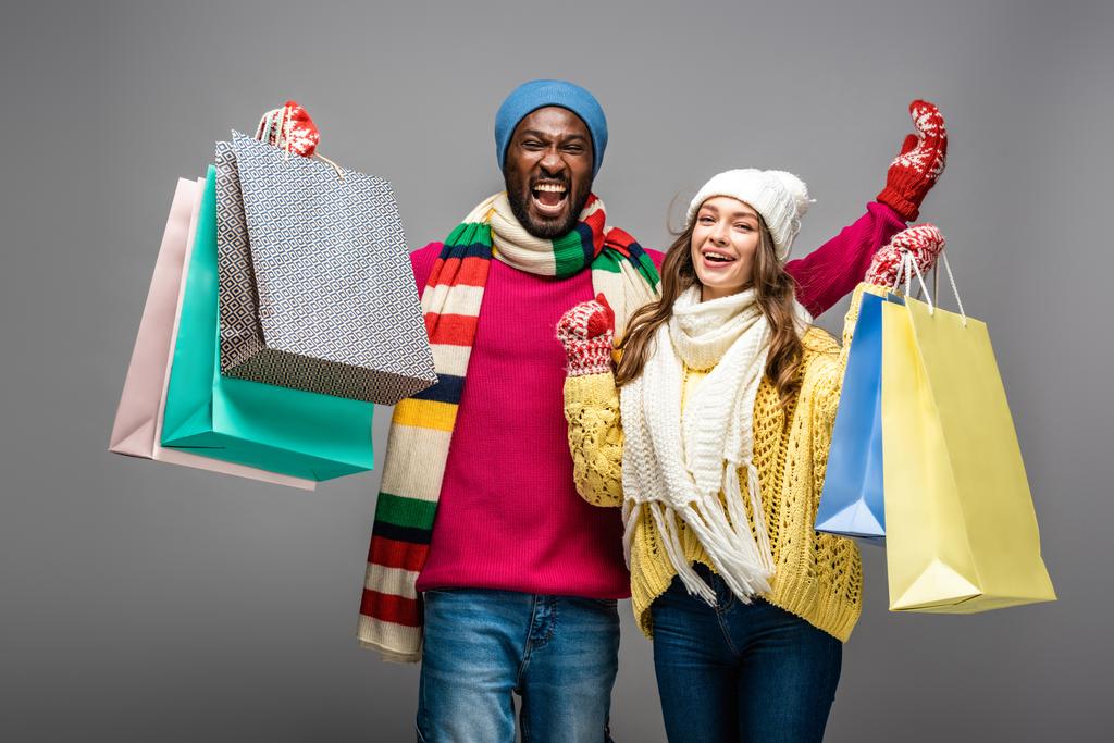 feliz casal interracial no inverno roupa segurando sacos de compras no fundo cinza
 - Foto, Imagem