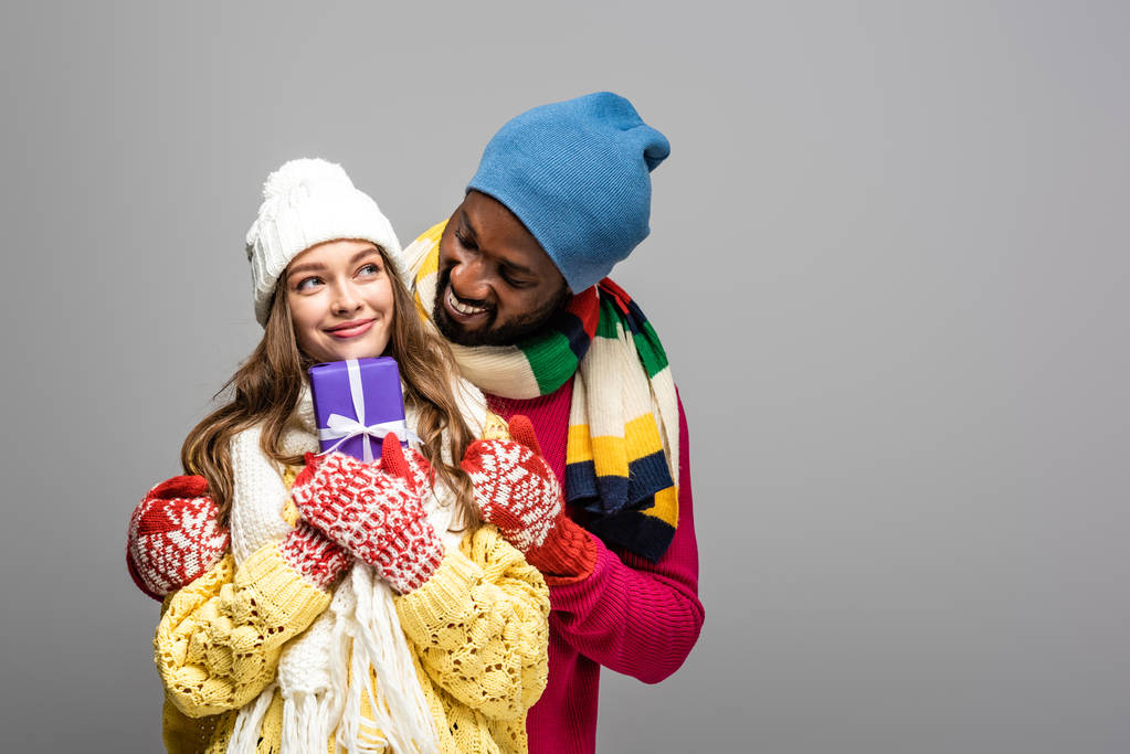 happy interracial couple in winter outfit holding present isolated on grey - Photo, Image
