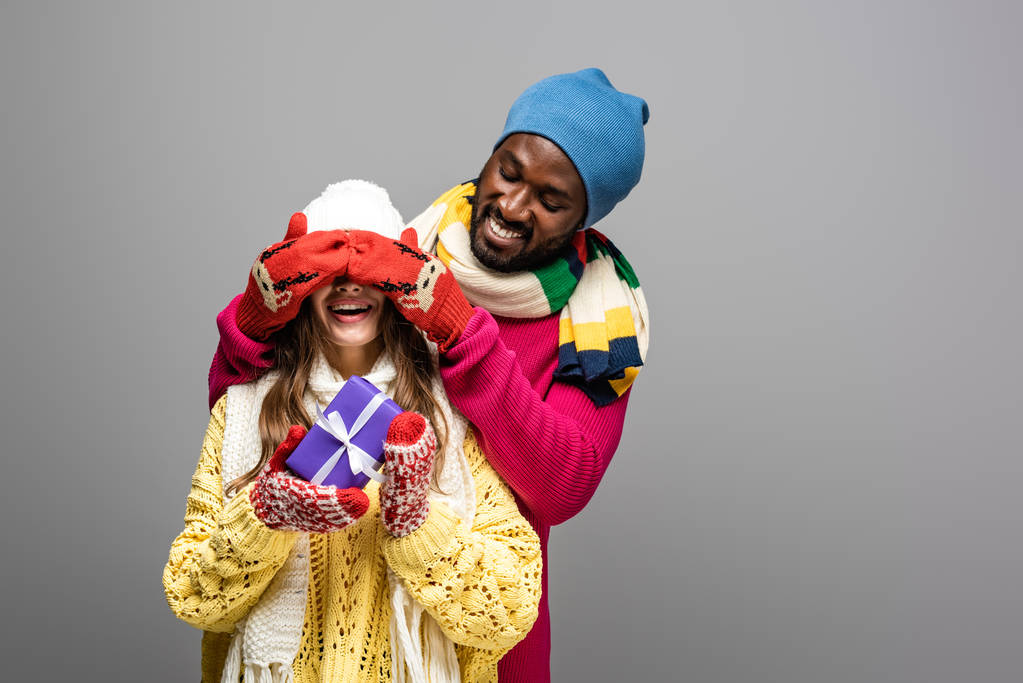 happy african american man in winter outfit covering eyes of girlfriend holding present isolated on grey - Photo, Image