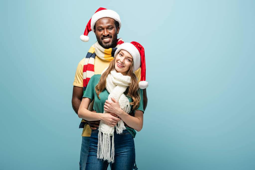 happy interracial couple in santa hats and scarves hugging isolated on blue - Photo, Image