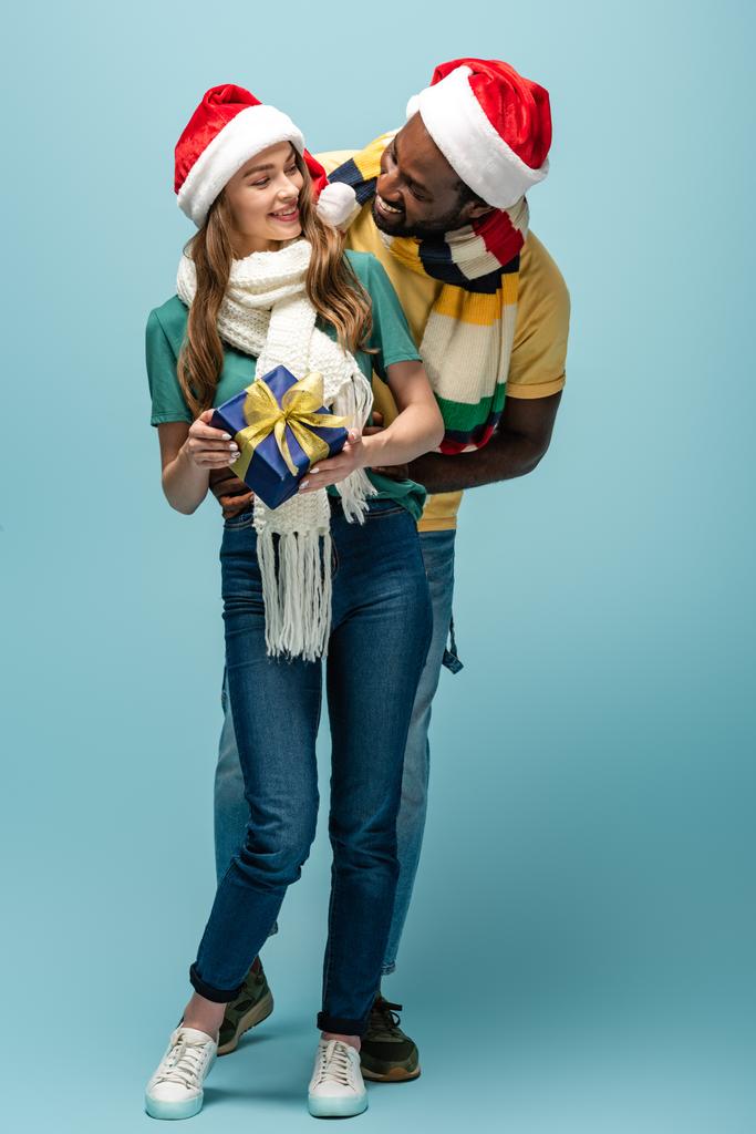 heureux homme afro-américain en santa chapeau et foulard cadeau cadeau à petite amie sur fond bleu
 - Photo, image