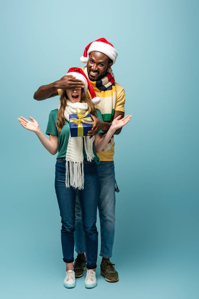 幸せなアフリカ系アメリカ人男性でサンタ・ハットとスカーフプレゼントに驚きのガールフレンドとカバーの目に青の背景 - 写真・画像