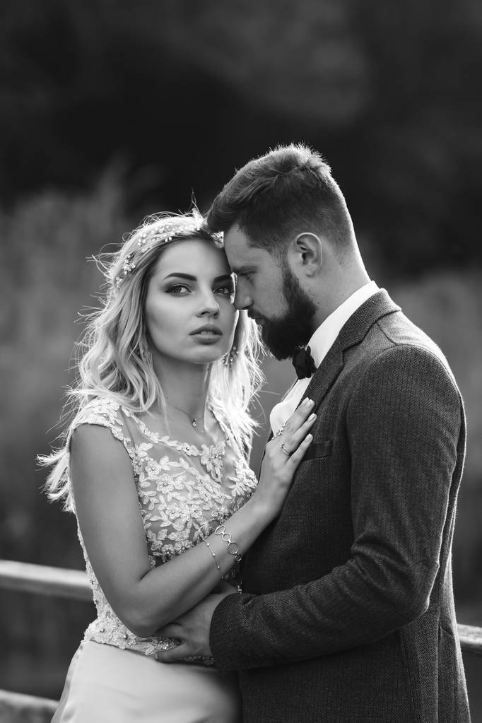 Fekete-fehér foto stílusos pár friss házasok pózol egy hídon az esküvő napján. Jóképű szakállas vőlegény csodálja és csókolja a csinos menyasszonyt. Együtt. Az ifjúság, a szerelem, a divat és az életmód fogalma. - Fotó, kép
