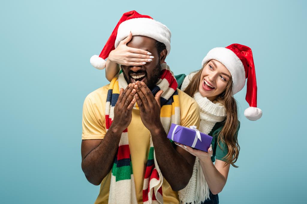 heureuse fille dans santa chapeau et écharpe couvrant les yeux sur afro-américain copain et cadeau isolé sur bleu
 - Photo, image
