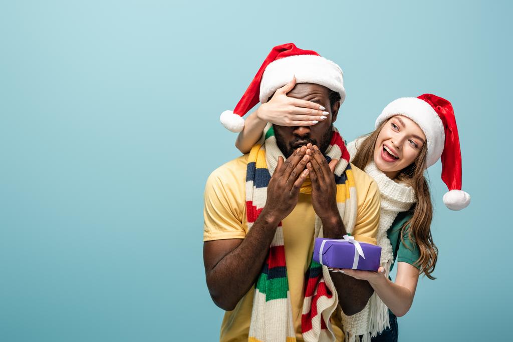 счастливая девушка в шляпе Санты и шарфе закрывая глаза на африканского американского бойфренда и подарка подарок изолирован на синий
 - Фото, изображение