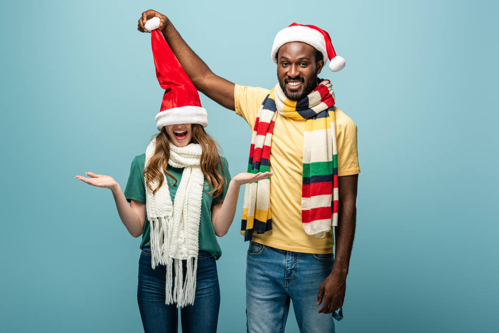 サンタの帽子とスカーフの幸せな異人種間のカップルは青で隔離され - 写真・画像