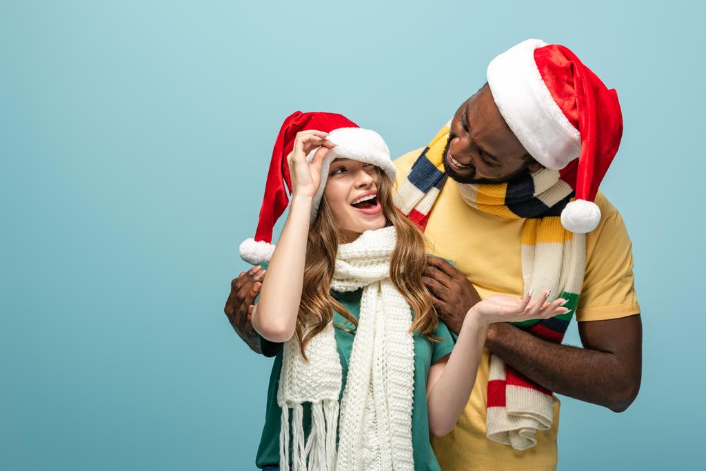 happy interracial couple in santa hats and scarves having fun isolated on blue - Photo, Image