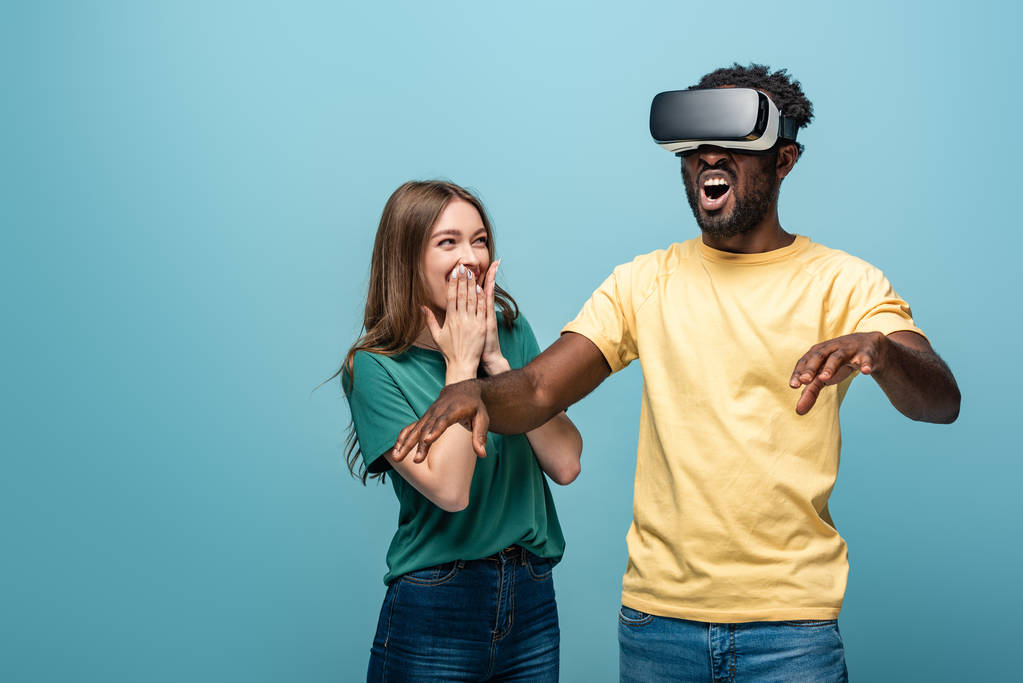 laughing girl standing near gesturing african american boyfriend in vr headset on blue background - Photo, Image