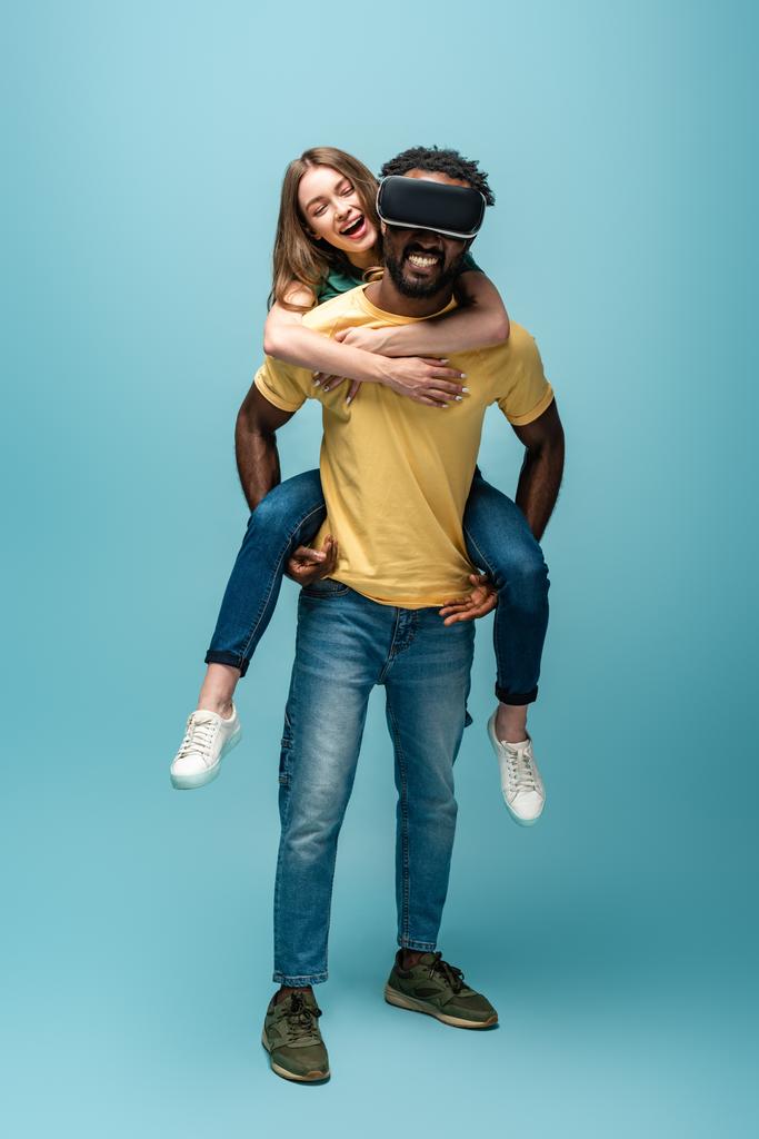 gelukkig meisje meeliften op afrikaanse amerikaanse vriend in vr headset op blauwe achtergrond - Foto, afbeelding