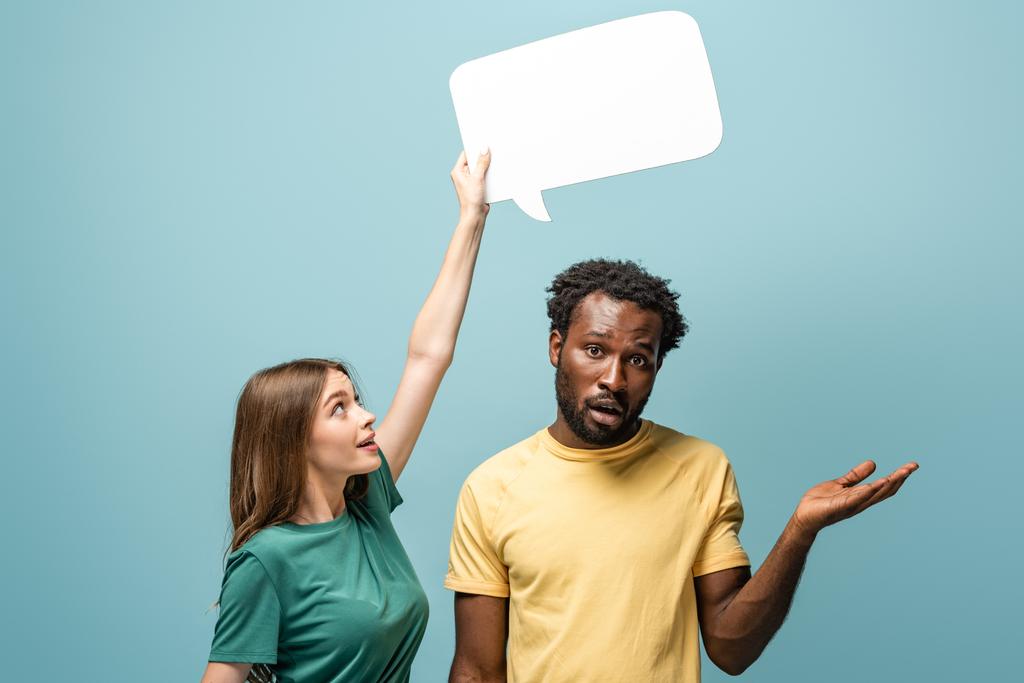 girl holding speech bubble above confused african american man showing shrug gesture on blue background - Photo, Image