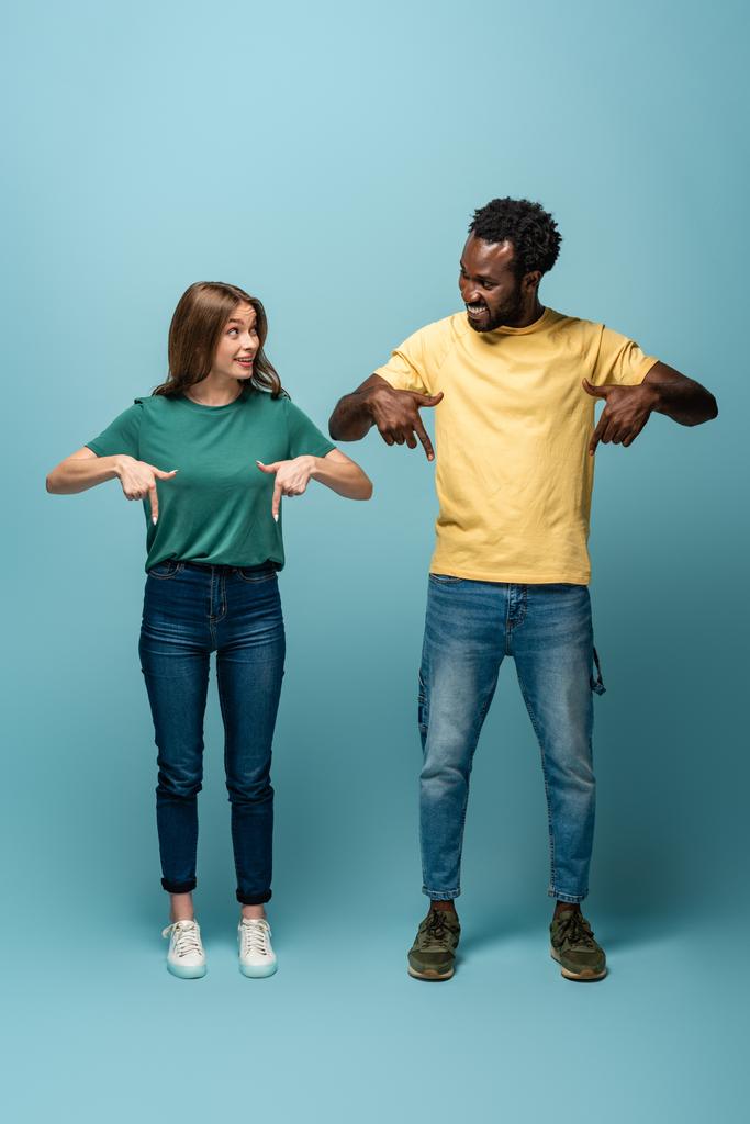 happy interracial couple pointing with fingers down on blue background - Photo, Image