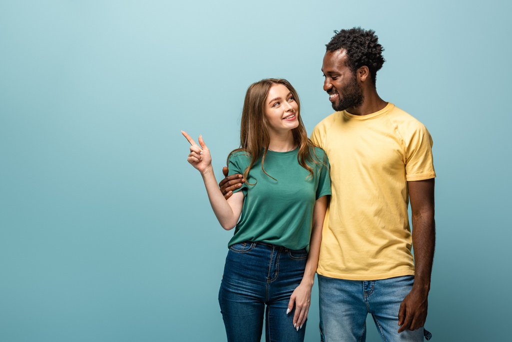 sonriente interracial pareja apuntando con el dedo lejos en azul fondo
 - Foto, imagen