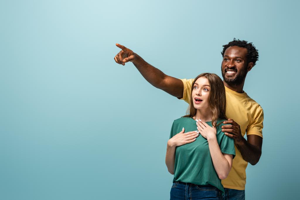 happy and surprised interracial couple pointing with finger away on blue background - Photo, Image
