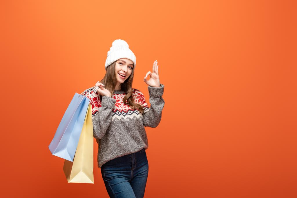счастливая девушка в зимнем наряде, держа в руках сумки с покупками и показывая знак ОК на оранжевом фоне
 - Фото, изображение