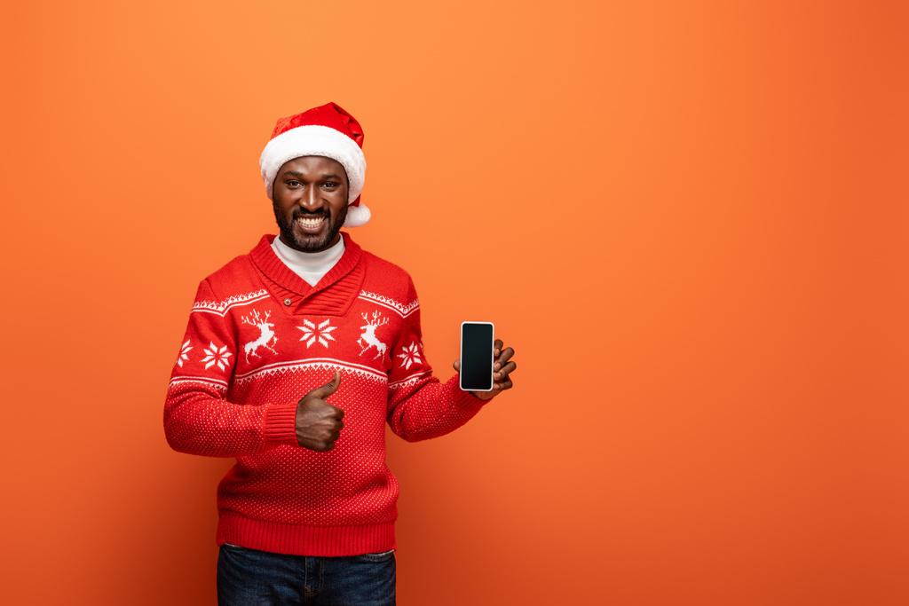 smiling african american man in santa hat and Christmas sweater presenting smartphone and showing thumb up on orange background - Photo, Image