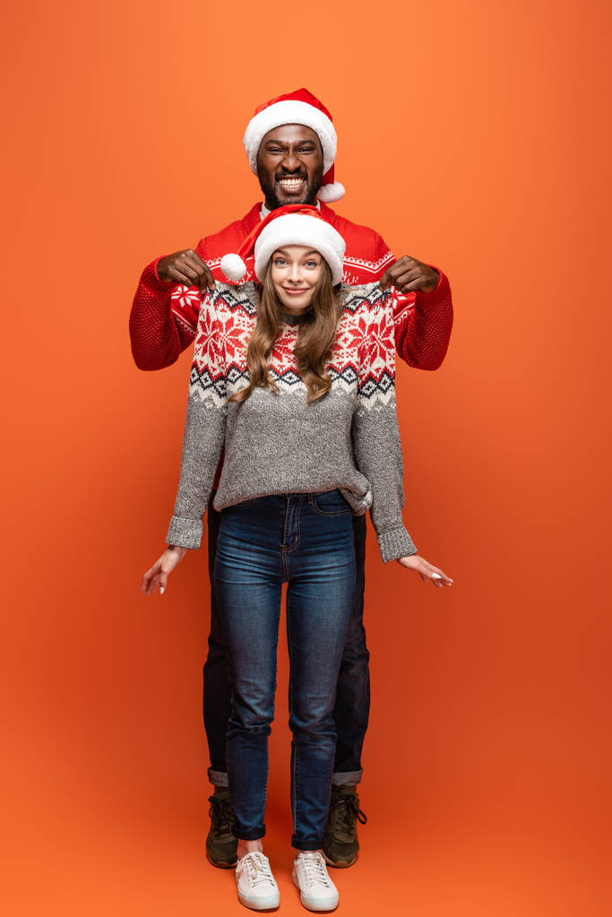 hombre afroamericano feliz levantando novia en suéter de Navidad sobre fondo naranja
 - Foto, imagen