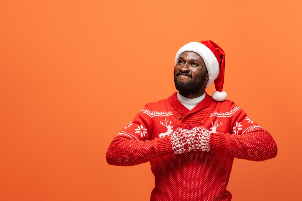 boze Afro-Amerikaanse man met kerstmuts, wanten en kersttrui op oranje achtergrond - Foto, afbeelding
