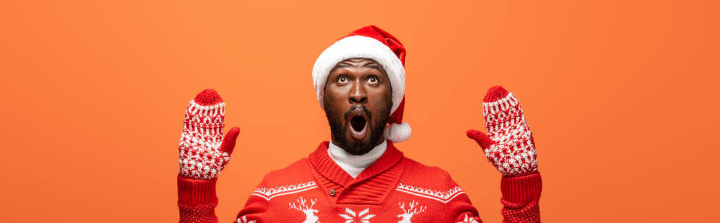 shocked african american man in santa hat, mittens and Christmas sweater isolated on orange, panoramic shot - Photo, Image
