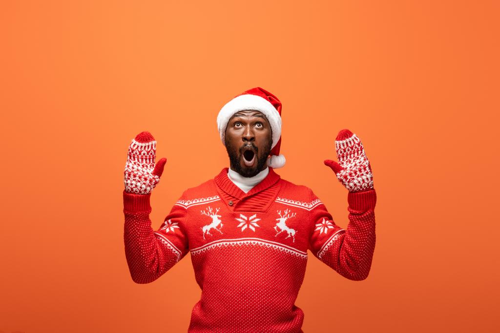 šokovaný africký Američan muž v Santa klobouku, palčáky a vánoční svetr na oranžovém pozadí - Fotografie, Obrázek