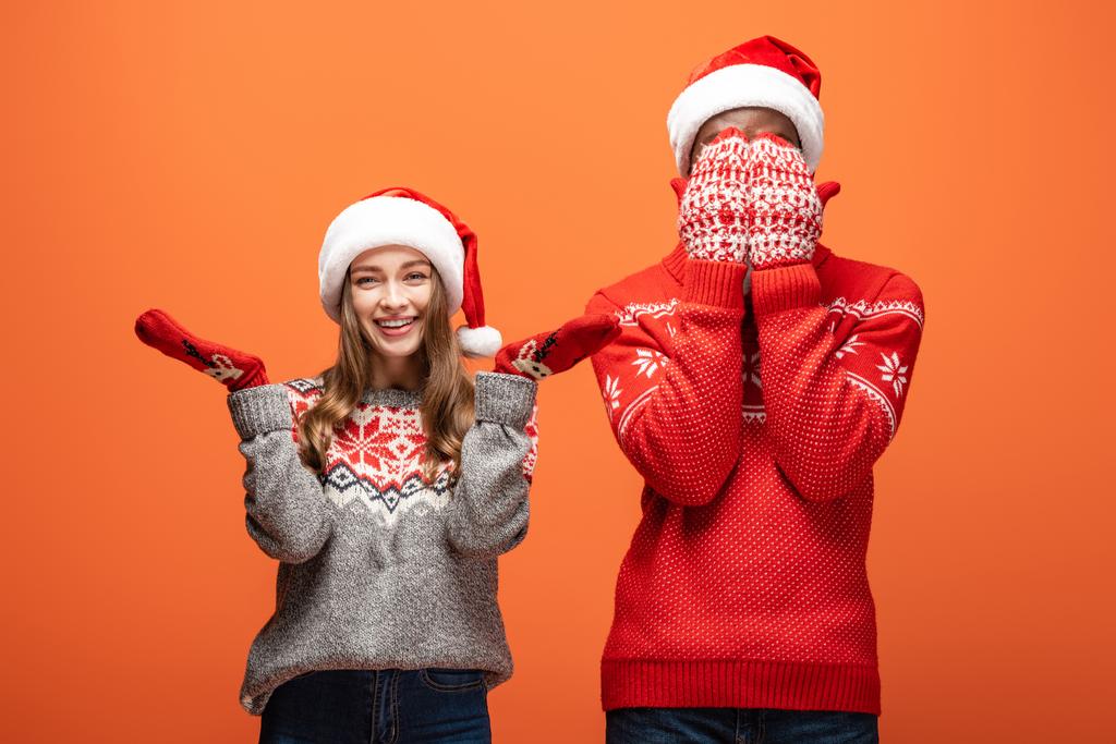Африканский американец, прикрывающий лицо руками счастливой девушки в рождественском свитере на оранжевом фоне
 - Фото, изображение